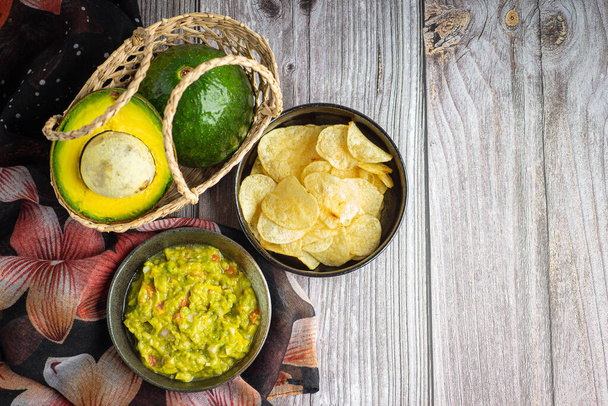 Čerstvý guacamole s bramborovými lupínky v misce a čerstvými avokádami v košíku na dřevěném stole. Mexické kuchyňské přísady. Horní pohled s místem pro kopírování. Koncept zdravého ovoce - Fotografie, Obrázek