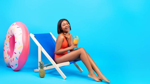 Full length of black woman drinking tropical cocktail in lounge chair, wearing swimsuit and laughing on blue background - Photo, Image