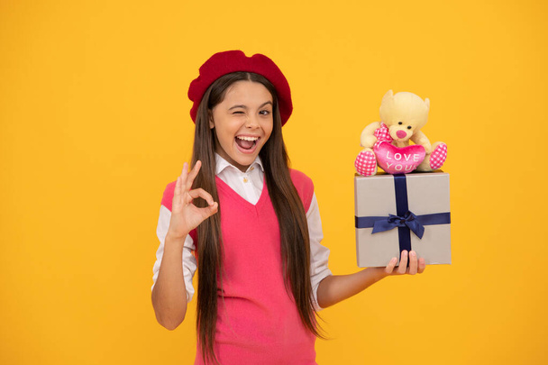 Ευτυχισμένος έφηβος κορίτσι στα γαλλικά μπερέ κρατήσει δώρο παιχνίδι και κουτί δώρου δείχνουν ok χειρονομία, παρουσιάζουν. - Φωτογραφία, εικόνα