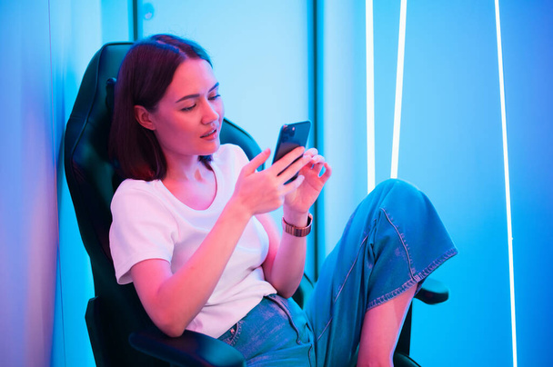 Dziewczyna relaksująca się grając w gry na telefonie komórkowym i siedząc na krześle do gier. Pokój z kolorowym świetle neonu. - Zdjęcie, obraz
