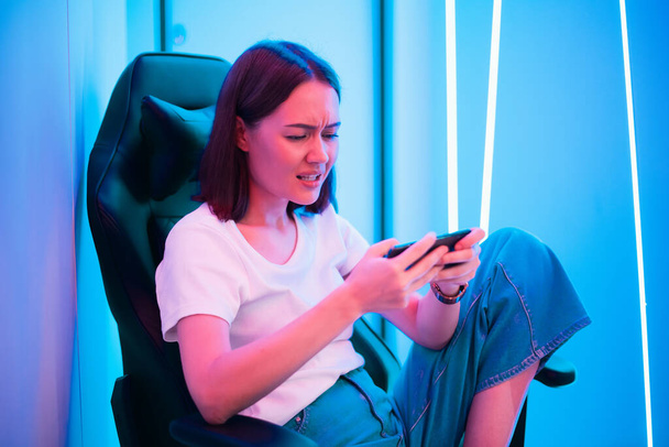 Beautiful young woman in white shirt with jeans uses smartphone in multicolored neon pink light at home at night. - Photo, Image
