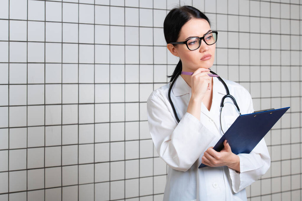 Jovem médica bonita em casaco cirúrgico branco com estetoscópio preto e suporte de papel azul em mãos em pé no consultório médico - Foto, Imagem