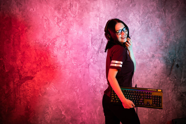 Female esports gamer posing with a gaming gear in neon light. Streamer girl standing with a gaming keyboard. - Photo, Image