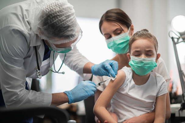 Doświadczony pediatra szczepi małą dziewczynkę, matka trzyma dziecko w ramionach, dziewczyna się boi - Zdjęcie, obraz