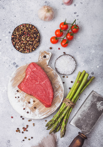 Rauwe biefstuk biefstuk op ronde snijplank met tomaten, knoflook en aspergepunten en vleesbijl op lichte keukentafel achtergrond. - Foto, afbeelding