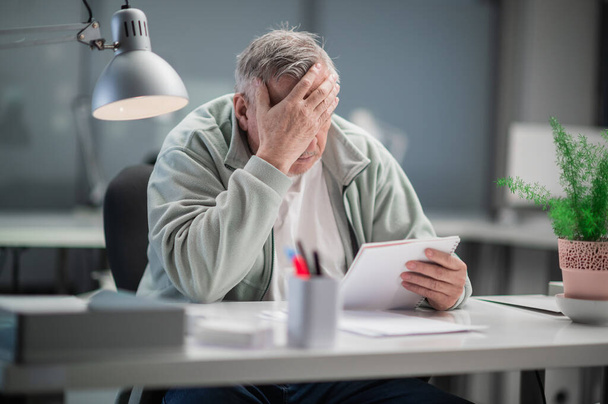 At the end of the working day, an elderly office worker felt a headache, he sits at his desk and holds his head - Photo, Image
