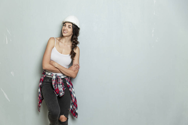 Smiling young woman in a white construction helmet against the background of a gray wall. Brunette in jeans and a plaid shirt. Construction, renovation and design. Space for text. - Φωτογραφία, εικόνα