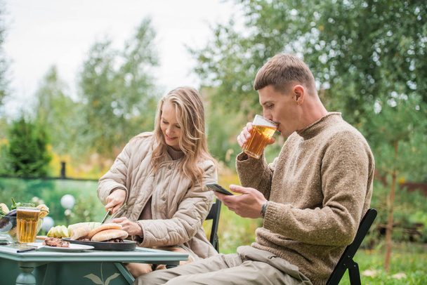 Ein Mann und eine junge Frau machten ein Picknick unter freiem Himmel im Hof eines Landhauses. Chillen am Tisch mit dem Smartphone in der Hand. - Foto, Bild