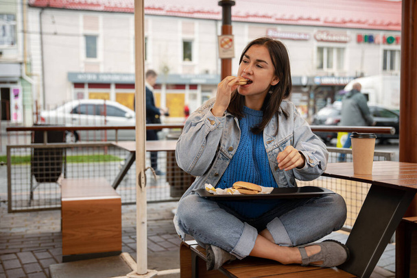 カジュアルなスタイルの魅力的な女の子は、カフェの夏のテラスに座ってコーヒーとバーガーを食べます. - 写真・画像