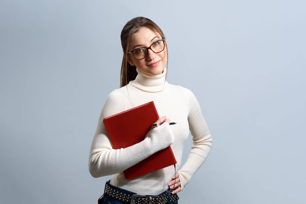 Portrait of a young woman with notepad and pen. Girl student with glasses on an isolated gray background. - Фото, изображение