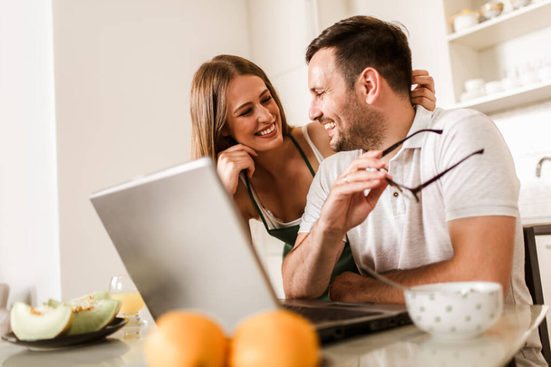 Νεαρό ζευγάρι περνούν χρόνο μαζί στο σπίτι στην κουζίνα χρησιμοποιώντας το laptop. - Φωτογραφία, εικόνα