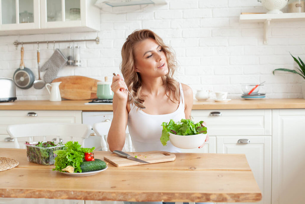 Jonge fitte vrouw bereidt en eet groentesalade in haar keuken. Gezond leven en gezond eten concept  - Foto, afbeelding