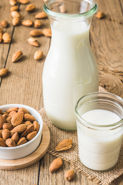Glass and pitcher with vegan almond milk, almond kernels and whole almonds on an old rustic wooden table, vertical - Photo, Image