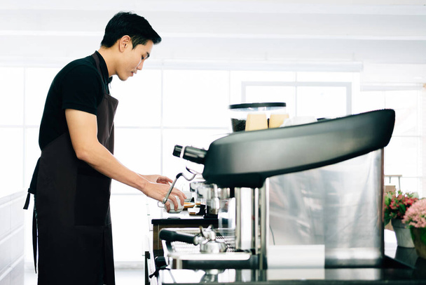 Νεαρό ασιατικό αρσενικό φορώντας ποδιά προετοιμασία φλιτζάνι καφέ και στέκεται μέσα στον πάγκο του καφέ με τη μηχανή του καφέ πριν από την παρασκευή καφέ. Τρόπος ζωής των επιχειρήσεων και των ανθρώπων κατά τη διάρκεια της πανδημίας Covid-19. - Φωτογραφία, εικόνα