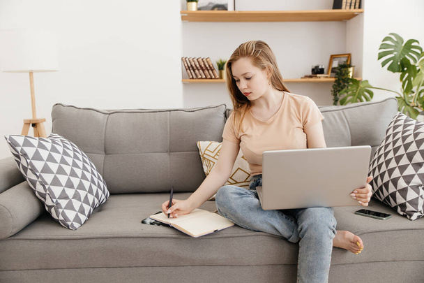 girl sitting on the couch works using a laptop and makes notes in a notebook - Foto, imagen
