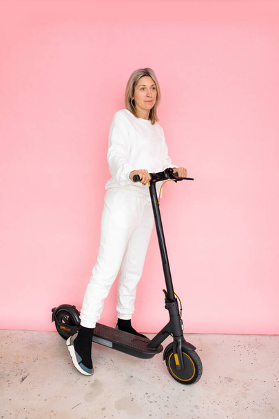 Одна жінка на електриці штовхає скутер на рожевий фон, студійний знімок, фотографія повної довжини
 - Фото, зображення