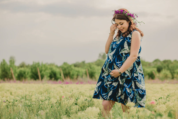 Mutterschaft und Schwangerschaft. Junge schwangere Frau im Blumenkranz berührt ihren großen Bauch auf einem Feld voller wilder Blumen und fühlt sich glücklich - Foto, Bild