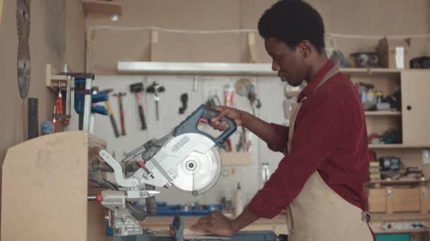 Tiro medio de carpintero africano masculino usando delantal usando sierra circular automática mientras trabaja en taller de carpintería - Imágenes, Vídeo
