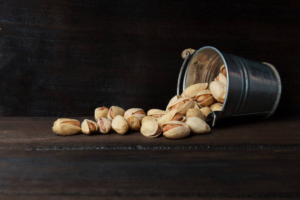 a bucket of pistachios fell on a dark wooden background. salted pistachios scattered in a mini bucket - Foto, Imagen