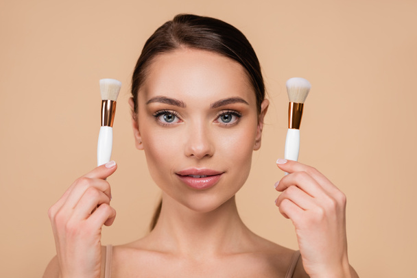 front view of young woman with natural makeup holding cosmetic brushes isolated on beige - Foto, Imagem