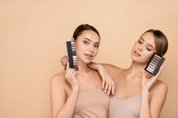 young woman leaning on shoulder of friend holding eye shadows palette isolated on beige - Fotoğraf, Görsel
