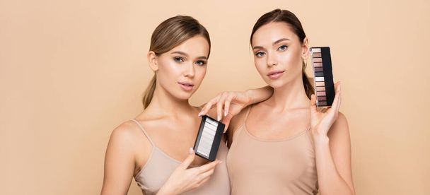 young women with natural visage holding eye shadows and highlighter palettes isolated on beige, banner - 写真・画像