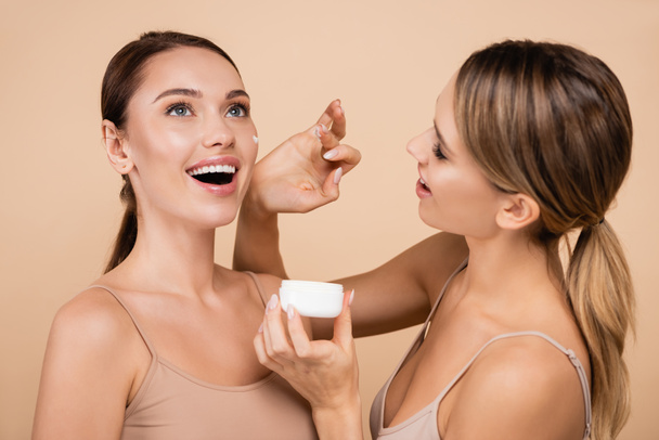 pretty woman applying face cream on excited friend isolated on beige - Photo, Image