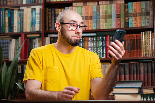 Viestintä. Kalju parrakas silmälasipäinen mies käyttää älypuhelinta. Taustalla on kirjasto ja hyllyt kirjoja. Sosiaalisten verkostojen, etäopetuksen ja karanteenin käsite. - Valokuva, kuva