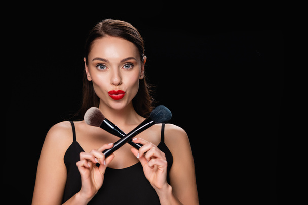 young woman with bright red lips posing with cosmetic brushes isolated on black - Fotoğraf, Görsel