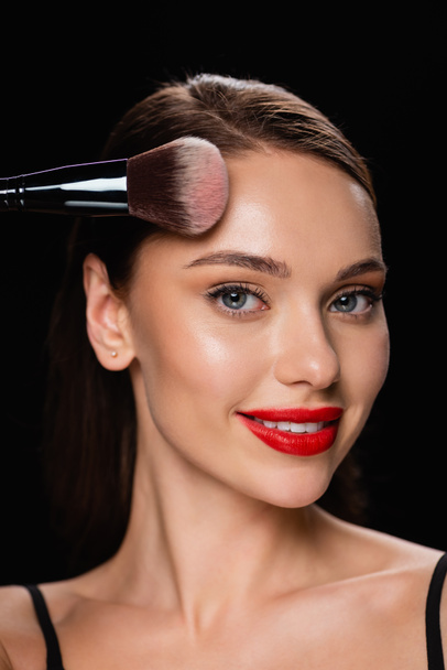 smiling woman looking at camera while powdering forehead isolated on black - Photo, image