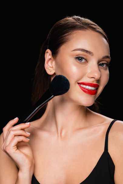 smiling woman powdering face with cosmetic brush isolated on black - Photo, image