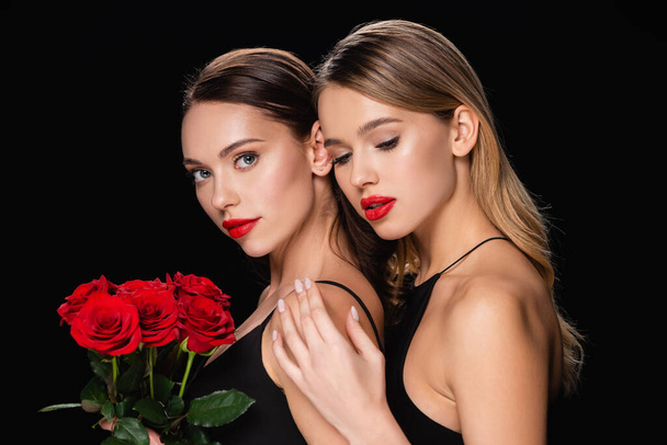 pretty woman touching shoulder of charming friend holding red roses isolated on black - Fotoğraf, Görsel