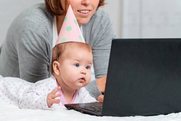 Portret van een baby met vakantiepet kijk naar de laptop. Moeder op de achtergrond. Sluit maar af. Concept van het gebruik van online videocommunicatie en internet. - Foto, afbeelding