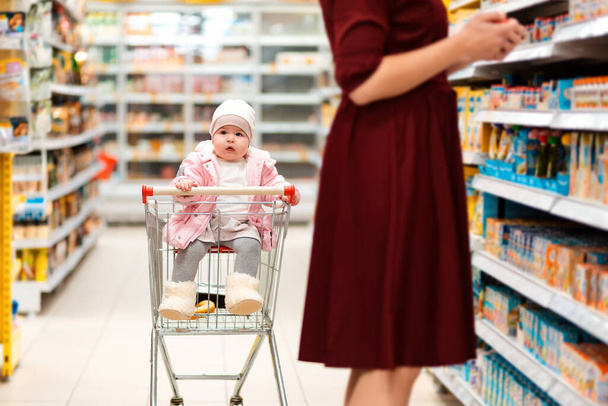 Ψώνια. Ένα νήπιο κάθεται σε ένα καρότσι για ψώνια και κοιτάζει τριγύρω ενώ η μητέρα του διαλέγει τα ψώνια. Οικογενειακά ψώνια στο σούπερ μάρκετ. - Φωτογραφία, εικόνα