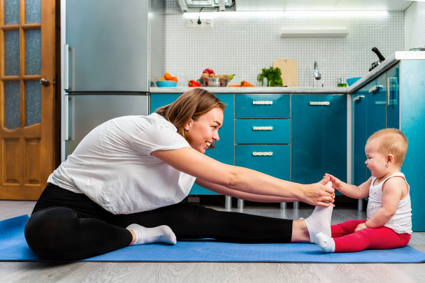 A smiling young mother and her baby are stretching on a mat in the kitchen. The concept of home sports training with children. - 写真・画像