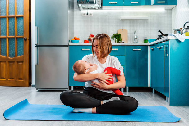 Sporttraining zu Hause. Eine junge, glückliche Mutter meditiert in der Küche, sitzt auf einer Sportmatte und hält das Baby in ihren Armen. Das Konzept von Yoga und Meditation. - Foto, Bild