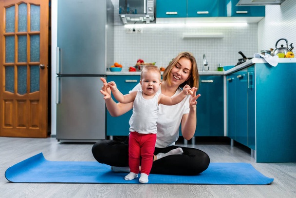A young mother meditates in the kitchen, sitting on a sports mat, teaching the baby to walk. The concept of yoga and baby's education. - Photo, Image