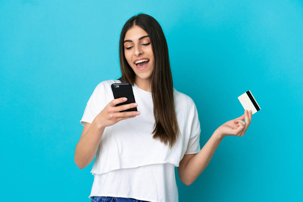 Молодая белая женщина, изолированная на синем фоне, покупает с мобильного с помощью кредитной карты - Фото, изображение