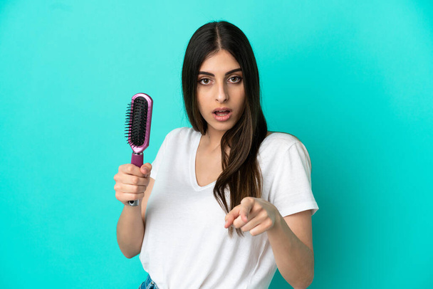 Young caucasian woman with hair comb isolated on blue background surprised and pointing front - Foto, Imagen