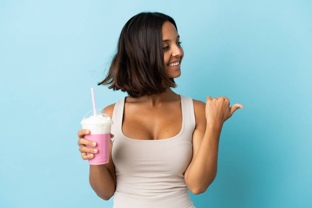 Mavi arka planda izole edilmiş çilekli milkshake ile genç bir kadın bir ürünü sunmak için yan tarafı işaret ediyor. - Fotoğraf, Görsel