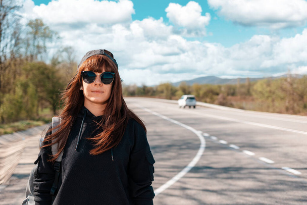 Portrait of caucasian young woman with backpack and sunglasses on the empty road. Copy space. The concept of local traveling and hitchhiking. - Photo, Image