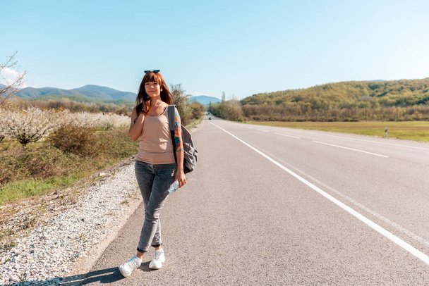 Hitchhiking. Smiling young woman with backpack and tattoed hand standing on the empty road. Road on the background. Copy space. The concept of local traveling. - Photo, Image