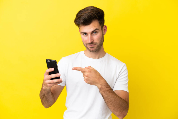 Young caucasian handsome man isolated on yellow background using mobile phone and pointing it - Fotó, kép