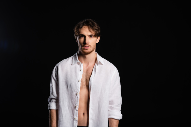 seductive man in unbuttoned white shirt looking at camera isolated on black - Foto, Imagen