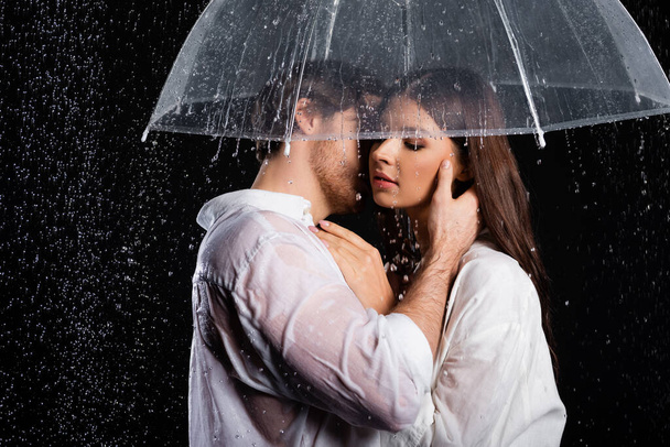 romantický mladý dospělý pár stojící v dešti s deštníkem a smyslně líbání na černém pozadí - Fotografie, Obrázek
