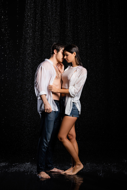 vue pleine longueur de couple sexy étreignant et embrassant dans des vêtements humides sous la pluie gouttes sur fond noir - Photo, image