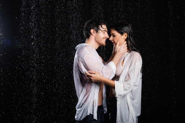 copain heureux et petite amie étreignant dans des vêtements humides sous la pluie sur fond noir - Photo, image
