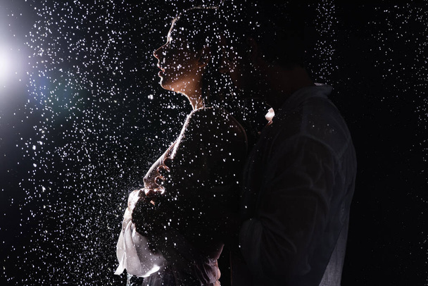 sexy pareja romántica apasionadamente abrazándose en gotas de lluvia sobre fondo negro con luz de fondo - Foto, imagen