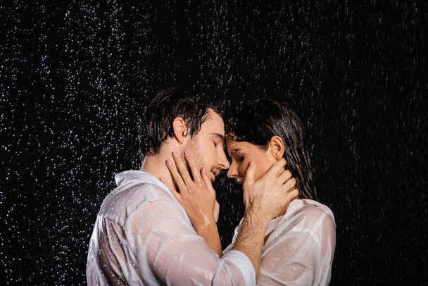 влажная страстная романтическая пара, держащаяся за руки рядом с лицами в дождевых капельках на черном фоне - Фото, изображение