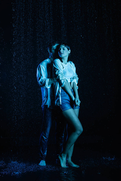 passionate man gently hugging wet sensual woman in water drops on black background with blue color filter - Photo, Image